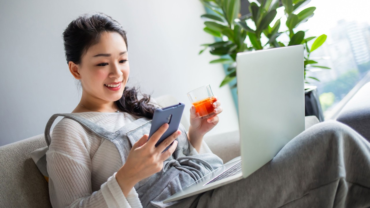 A lady is holding a drink and using phone with a laptop on her lap; imaged used for HSBC Taiwan foreign exchange page.